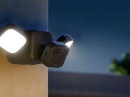 A Blink Outdoor 4 Floodlight Cam attached to the side of a home at dusk casts its 700 lumens of light into the yard.