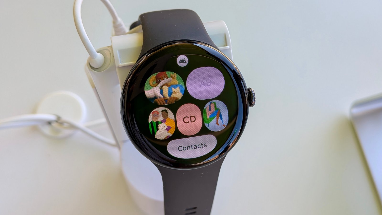 The Contacts Tile on Wear OS 6 on the Pixel Watch 3, showing different contact icons for individual numbers and an All Contacts button on the bottom edge.