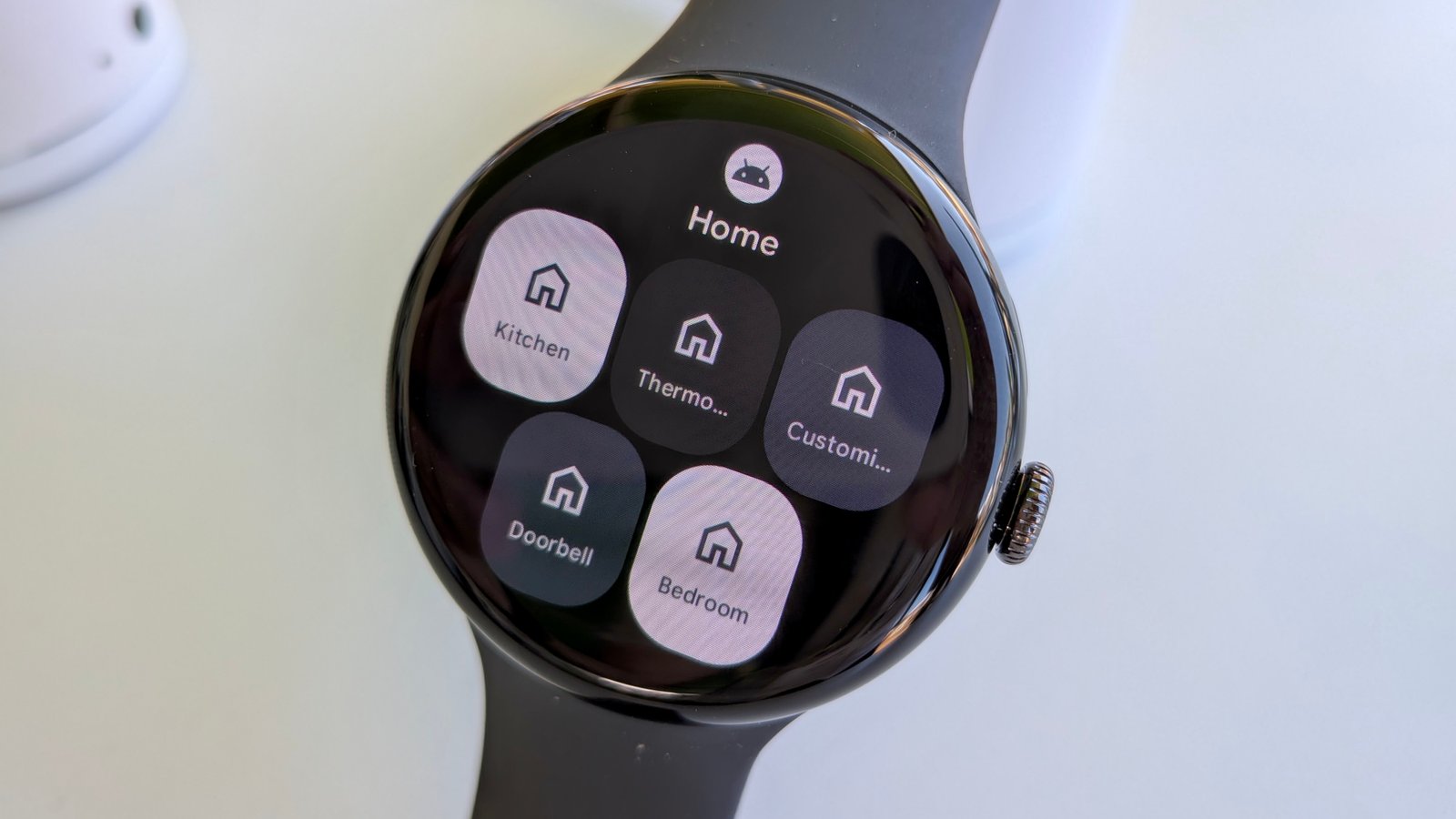 The new Google Home tile on Wear OS 6 on the Pixel Watch 3, showing options to check your Kitchen, Thermo, Custom, Doorbell, and Bedroom controls.