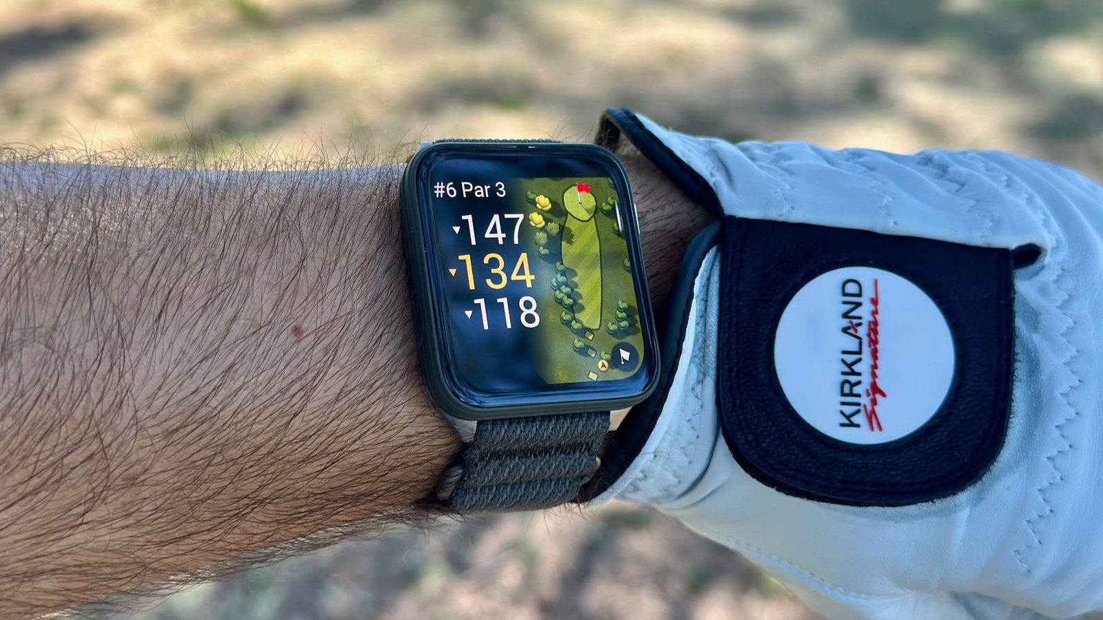 The Garmin Venu X1 on a wrist next to a golf glove, showing the yardage for the hole with fairway rough in the background.